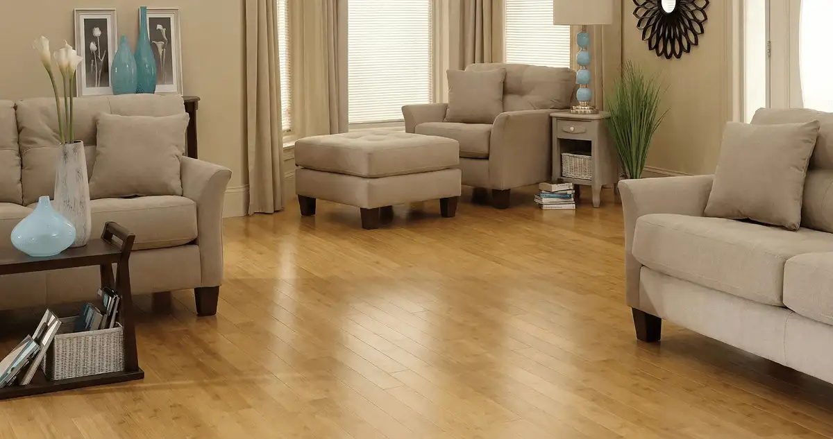 A living room with professionally installed bamboo flooring, beige sofas, a matching ottoman, a side table with a blue lamp, vases, framed pictures, and large windows with blinds and curtains in Tempe, AZ
